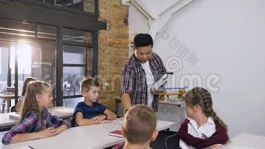 年轻的韩国教师走在课桌之间，把课本放在课桌上，而白种人学生则坐在课桌前